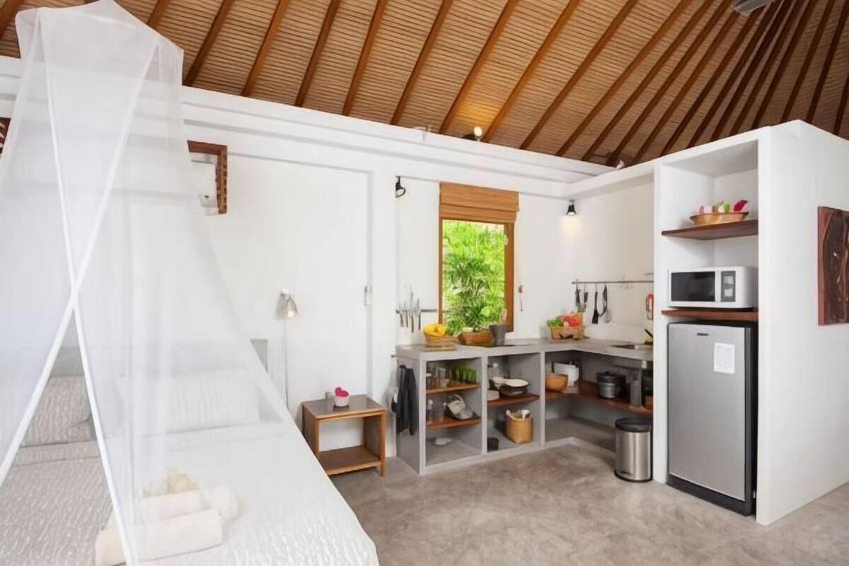 A bedroom with a canopy bed, a kitchenette with a refrigerator, and a window with a view of greenery.