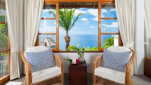 A room with two wicker chairs, a small table with a vase and flowers, and a large window with a view of the ocean.