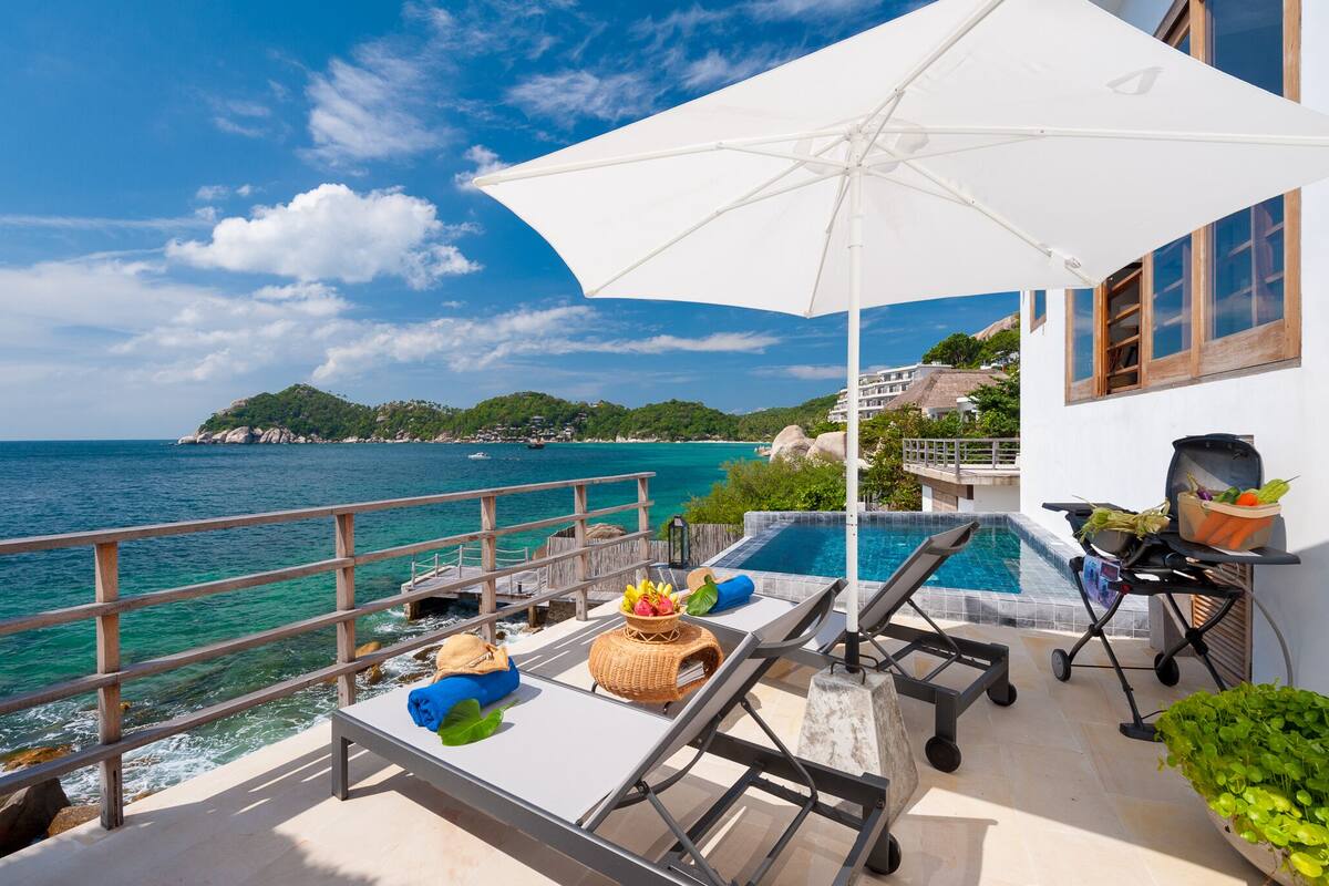 A poolside area with a view of the sea, a grill, lounge chairs, and a large umbrella.
