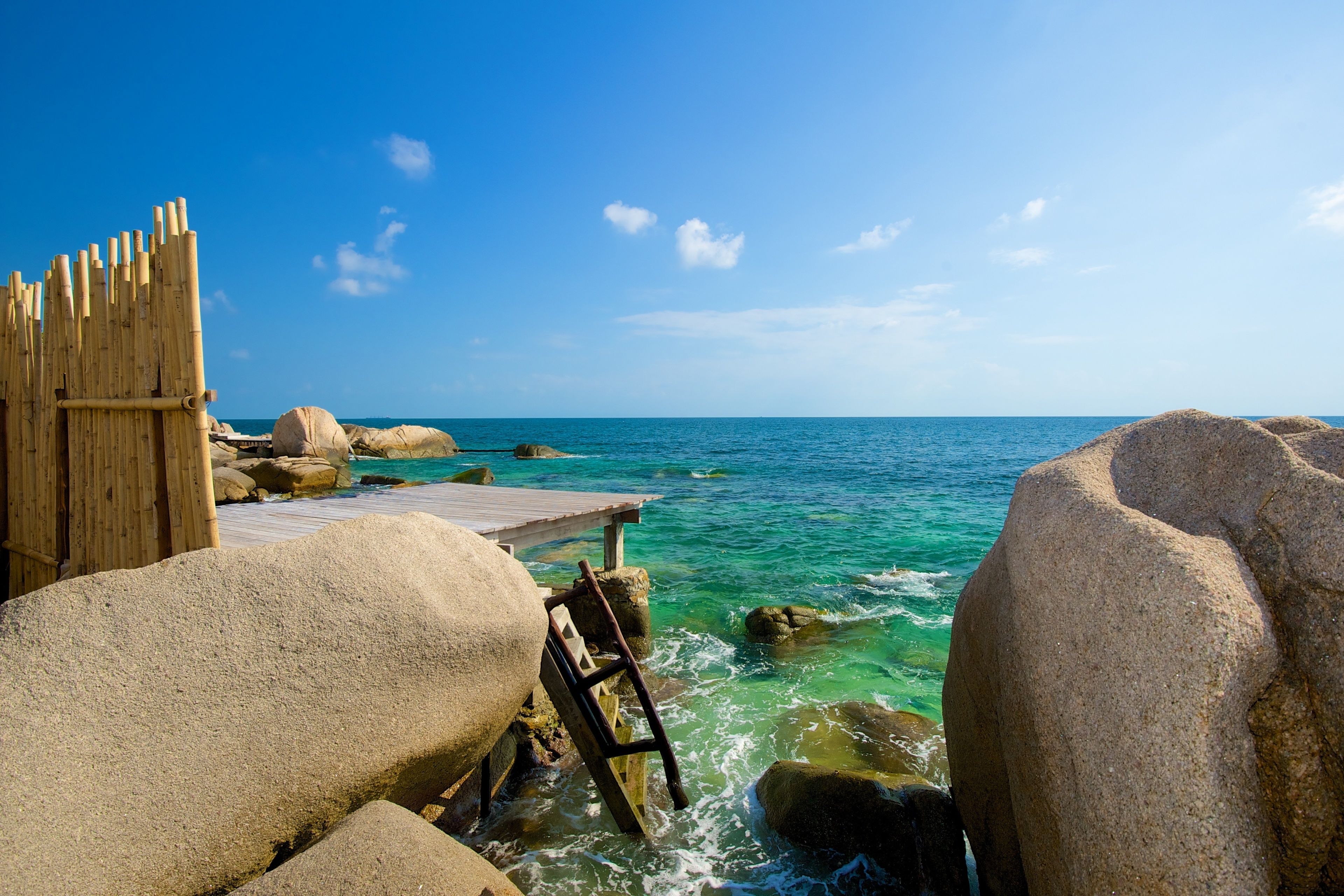 A wooden pier extending into a clear blue sea, surrounded by large rocks and a bamboo fence.