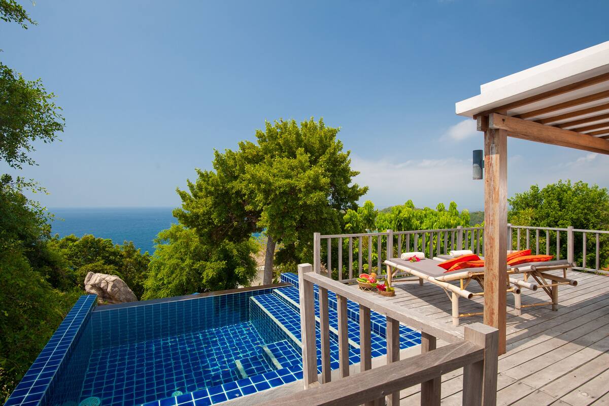 A rooftop infinity pool with wooden deck and lounge chairs, overlooking a scenic ocean view.