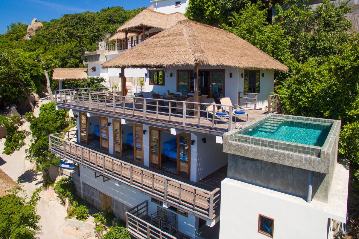 A two-story building with a thatched roof, a pool, and a balcony with seating.