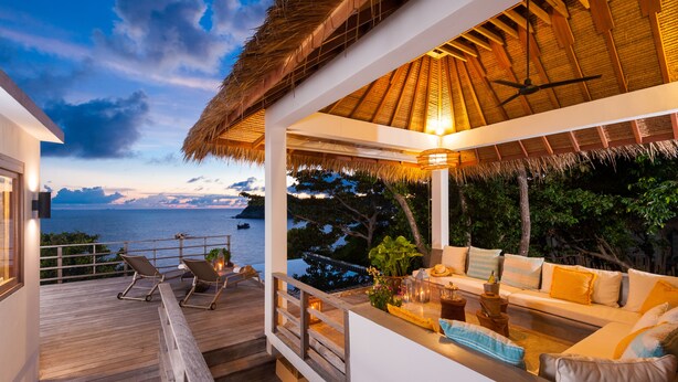 A covered outdoor seating area with a view of the sea, wooden deck, and a thatched roof structure.