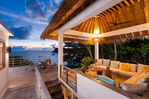 Un espace extérieur abrité, doté de sièges, offrant une vue sur la mer, avec une terrasse en bois et une structure surmontée d’un toit de chaume.