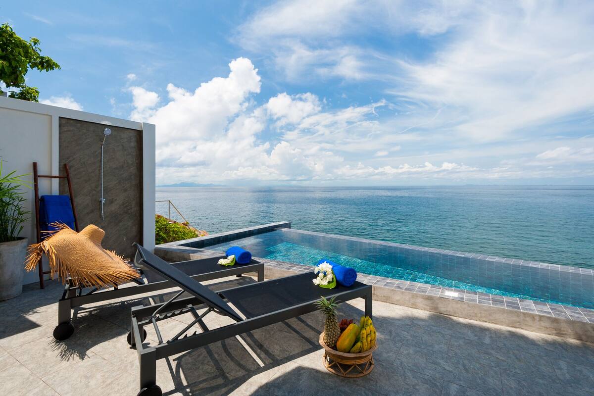 An infinity pool with a clear view of the ocean, lounge chairs, a small table with a fruit basket, and a potted plant.