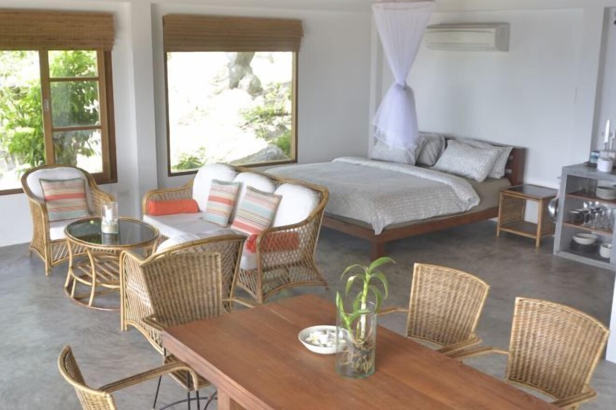 A bedroom with a bed, a dining table, wicker chairs, a small table with a vase, and a window with a view of greenery.