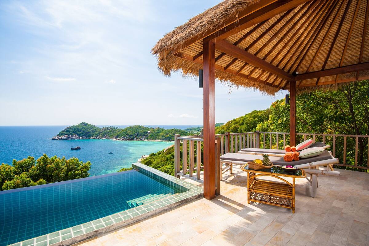 A poolside area with a thatched roof, lounge chairs, and a view of the sea and greenery.