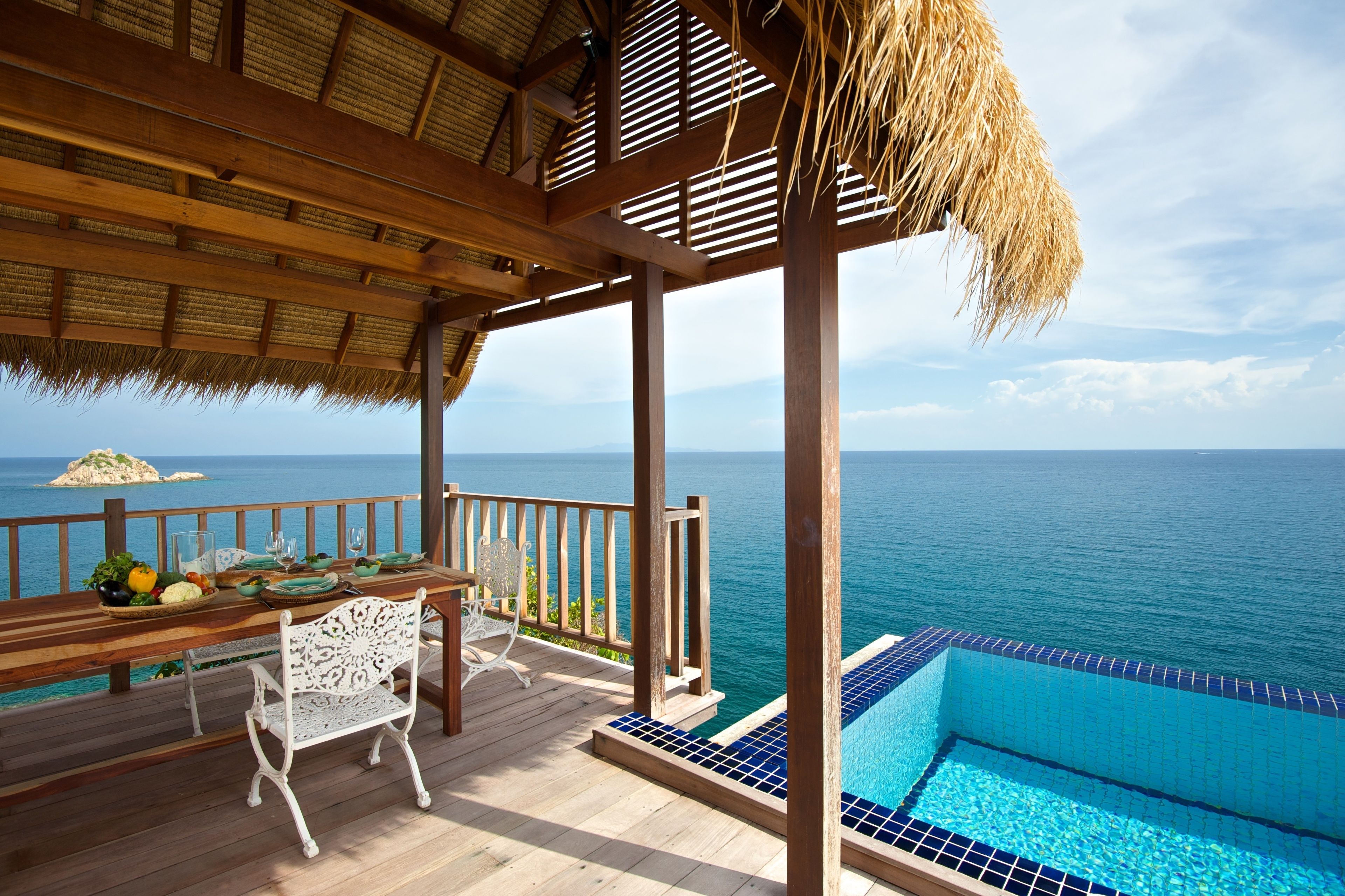 A wooden deck with a thatched roof overlooking a pool and the sea.