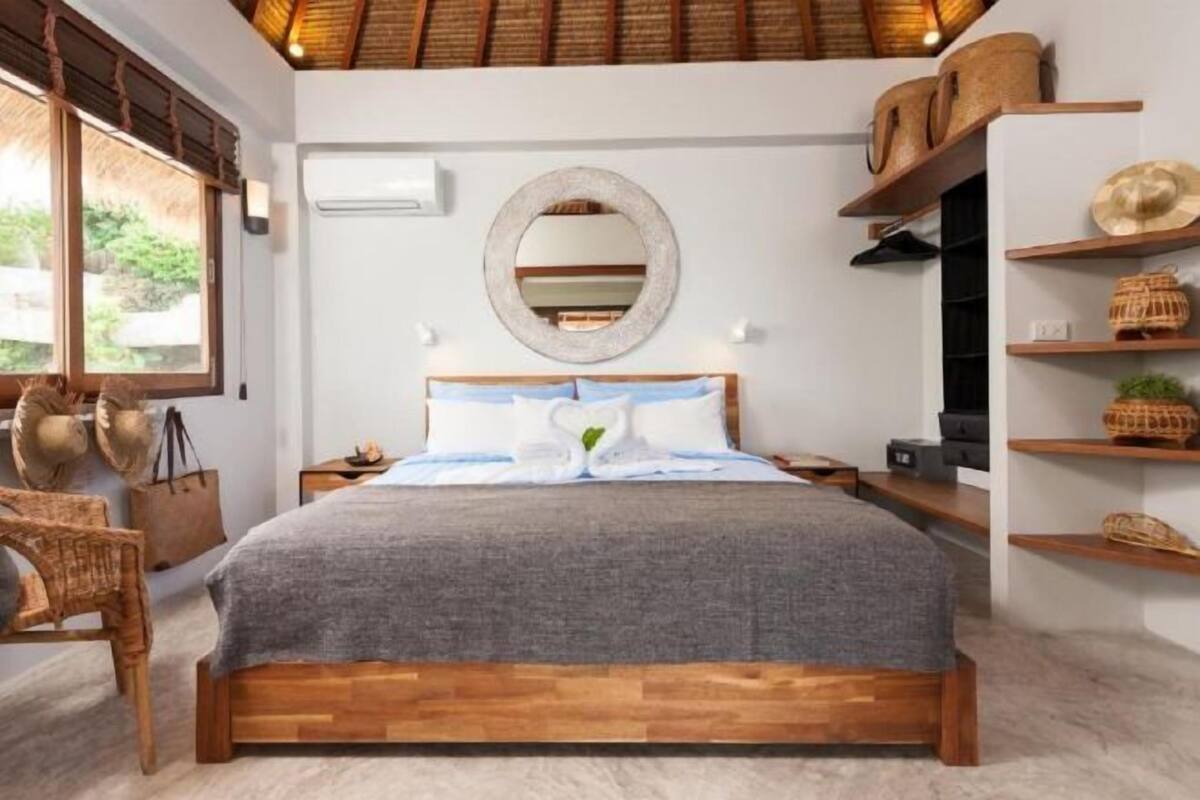 A bedroom with a wooden bed, a round mirror, wicker baskets, and a view of greenery outside.
