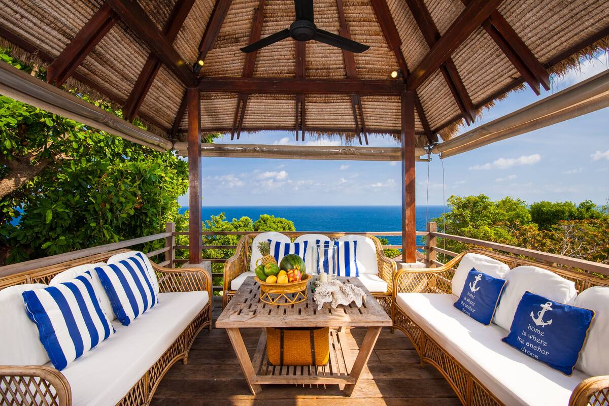A covered outdoor seating area with wicker furniture, a table with fruits and a fish, and a view of the ocean.
