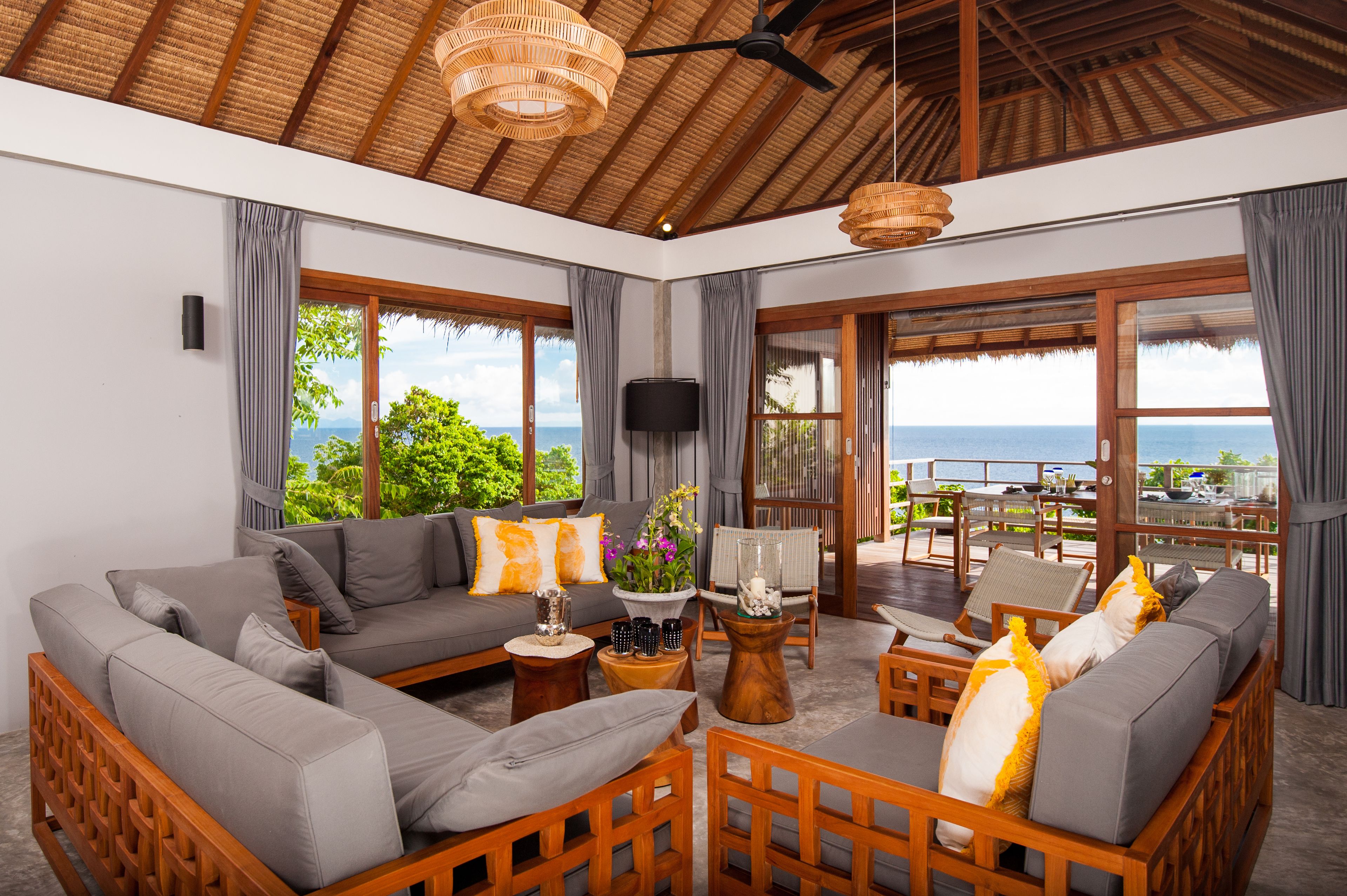 A spacious living area with a large sofa, wooden chairs, and a coffee table. There is a television mounted on the wall and a view of the ocean through large windows.