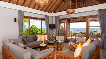 A spacious living area with a large sofa, wooden chairs, and a coffee table. There is a television mounted on the wall and a view of the ocean through large windows.