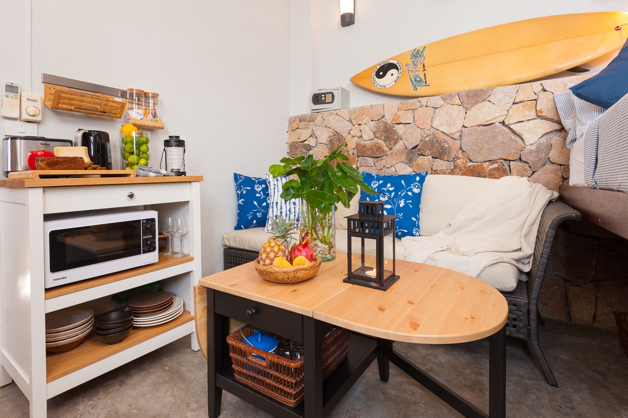 A compact living space with a kitchenette, a stone wall, a surfboard, and a cozy seating area.