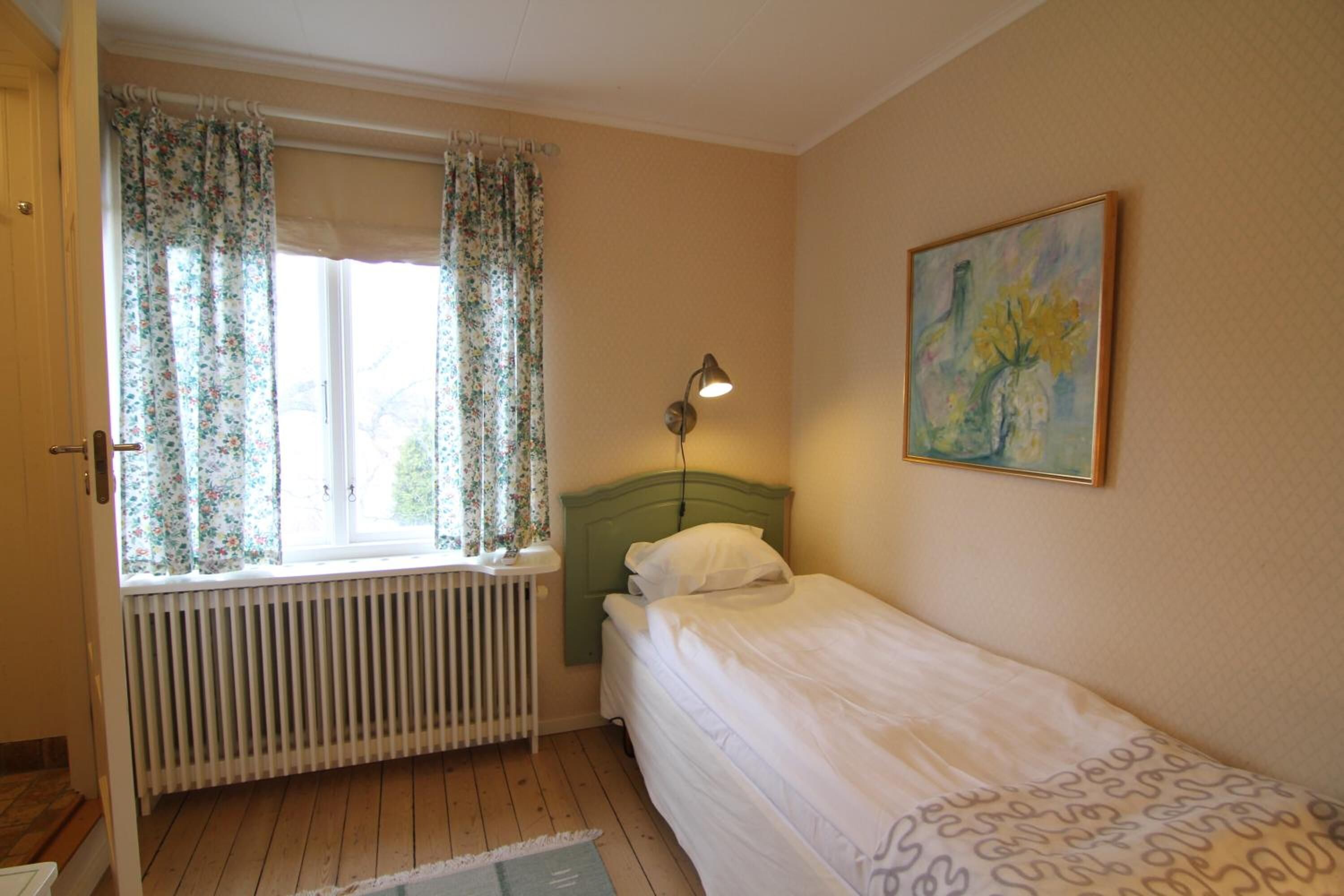A bedroom with a single bed, a radiator, a window with floral curtains, a bedside lamp, and a painting on the wall.