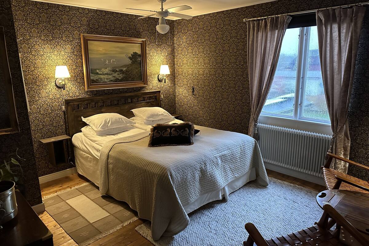 Un dormitorio con una cama, dos lámparas de noche, un ventilador de techo, una ventana con cortinas, un radiador y una mesa de madera.