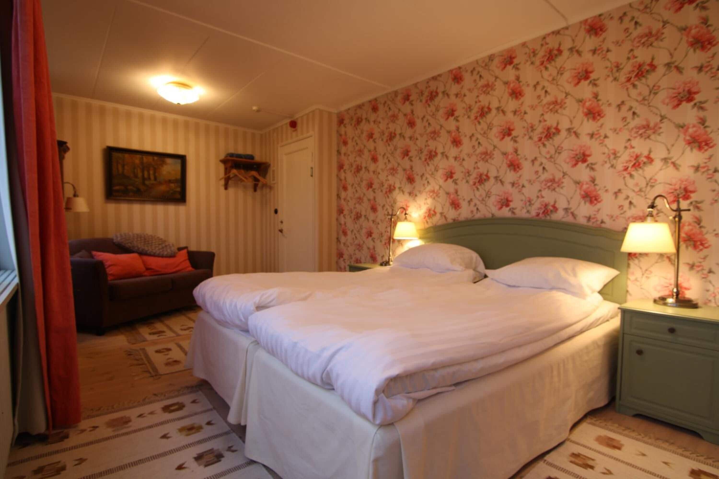 Un dormitorio con una cama doble, una mesita de noche con lámpara, un sofá con cojines, un cuadro en la pared y un estampado floral en el papel tapiz.