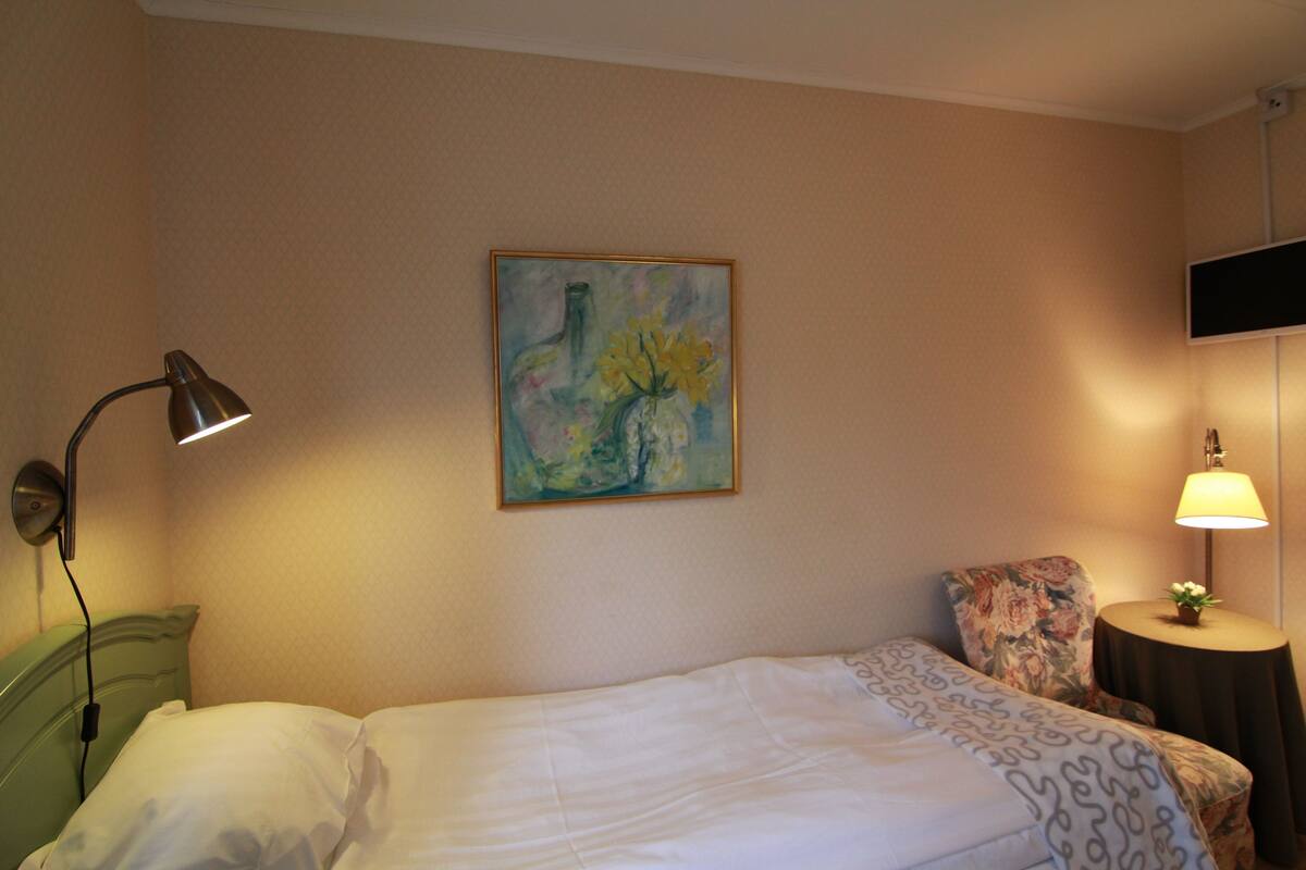 Habitación de hotel con una cama, un cuadro en la pared, una lámpara y una mesita con una planta.