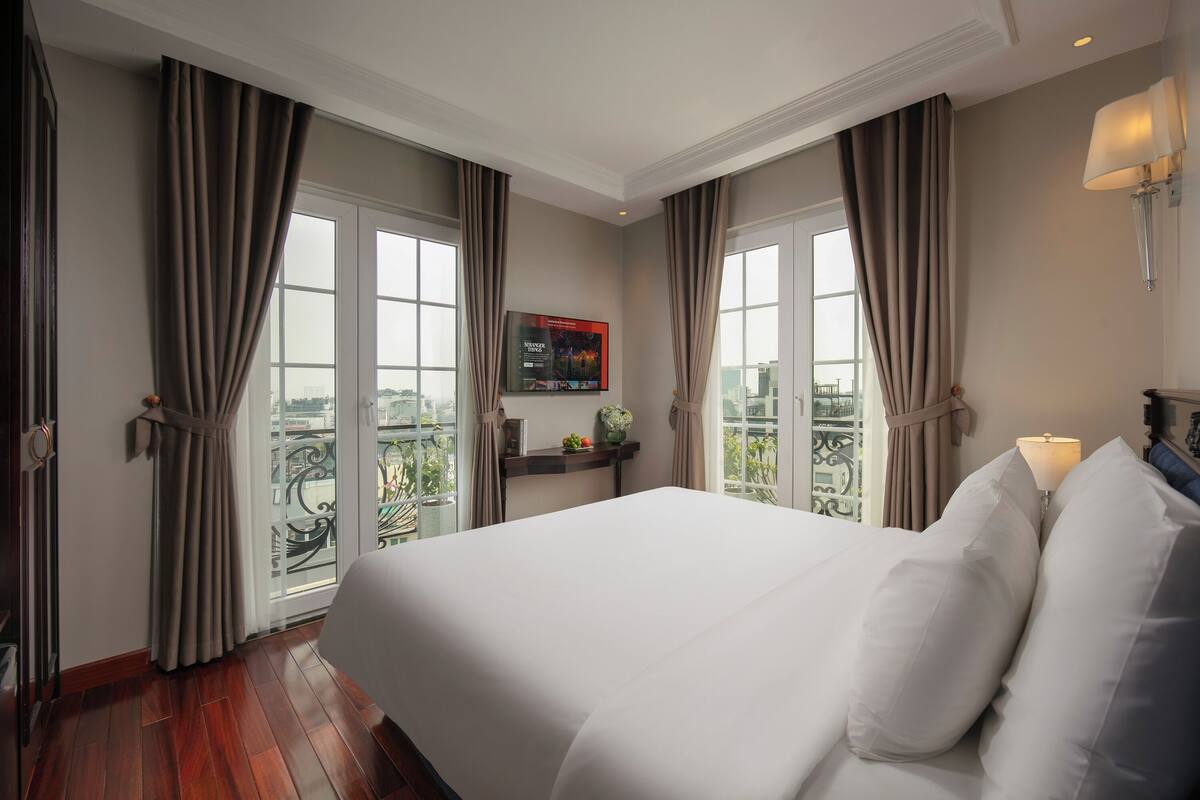 A hotel room with a large bed, a TV on the wall, and a balcony with curtains.