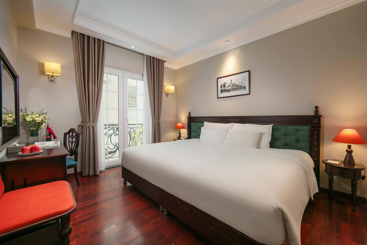 A hotel room with a large bed, a desk with a chair, a small table with a lamp, and a picture on the wall.