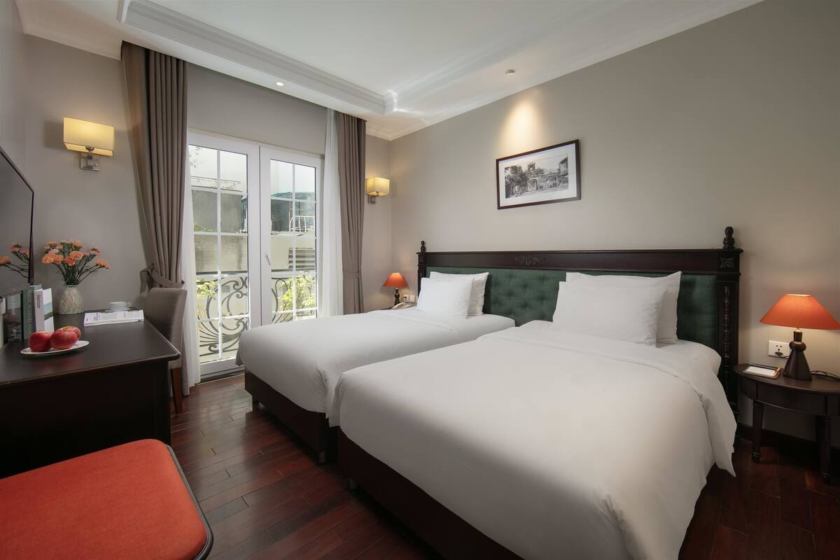 A hotel room with two beds, a desk with a chair, a small table with a lamp, and a balcony with a view.
