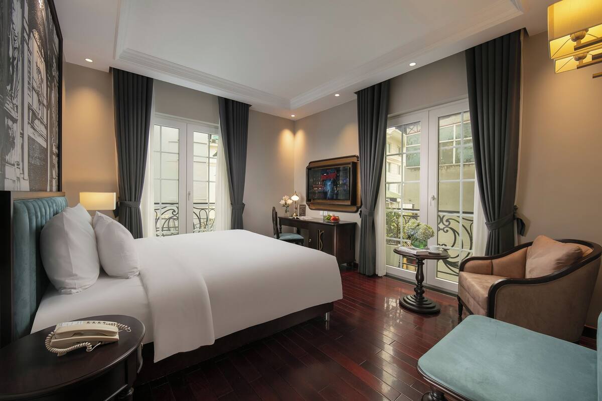 A hotel room with a large bed, a desk, a chair, a small table, and a balcony with a view.