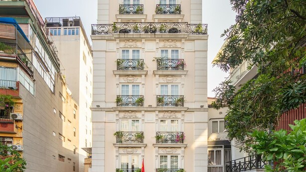 Ein mehrstöckiges Hotel im klassischen Stil, mit Balkonen, die mit Pflanzen bepflanzt sind, einem prunkvollen Eingang mit Säulen und einer Treppe, die zum Eingang führt.