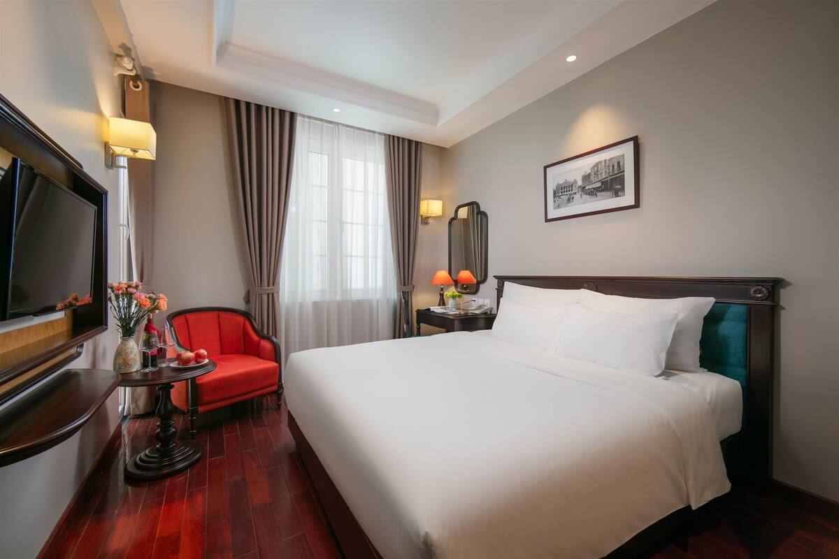 A hotel room with a large bed, a television, a red chair, a small table with flowers, and a framed picture on the wall.