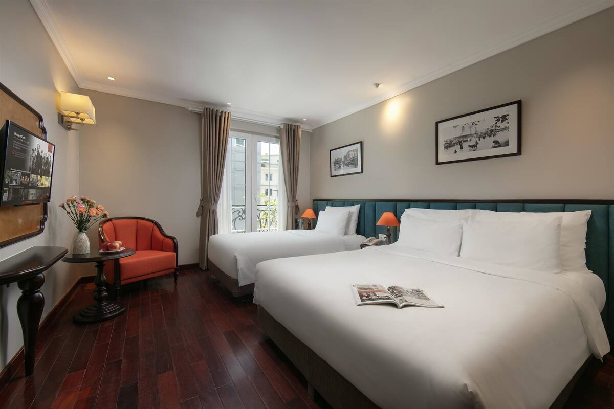 A hotel room with two beds, a small table with a vase of flowers, a chair, and a framed picture on the wall.