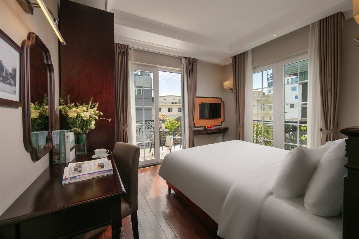 A hotel room with a large bed, a desk with a chair, a TV, and a balcony with a view of buildings.