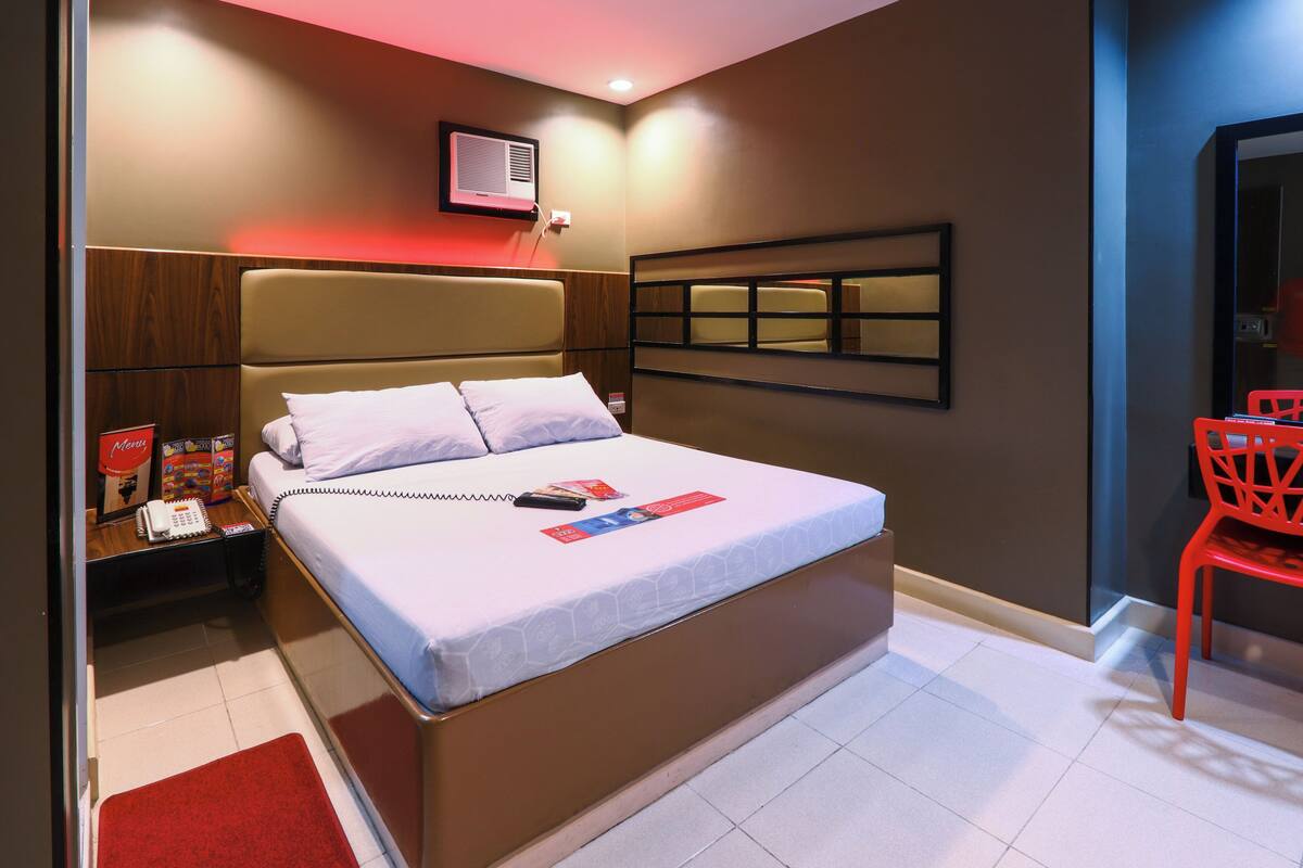 A modern hotel room with a large bed, a bedside table with a phone and magazines, a red chair, and a mirror on the wall.