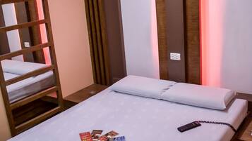 Una cama bien hecha con un control remoto y revistas, una litera de madera a la izquierda, y una pared con paneles de madera verticales y una luz roja.