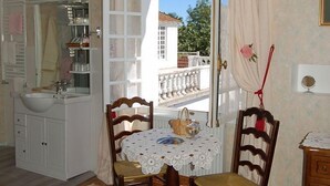 Quarto com toucador branco, mesa redonda com cadeiras, vista por uma porta aberta e varanda com guarda-corpo.