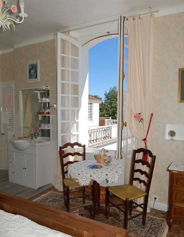 A room with a white vanity, a round table with chairs, a view through an open door, and a balcony with a railing.