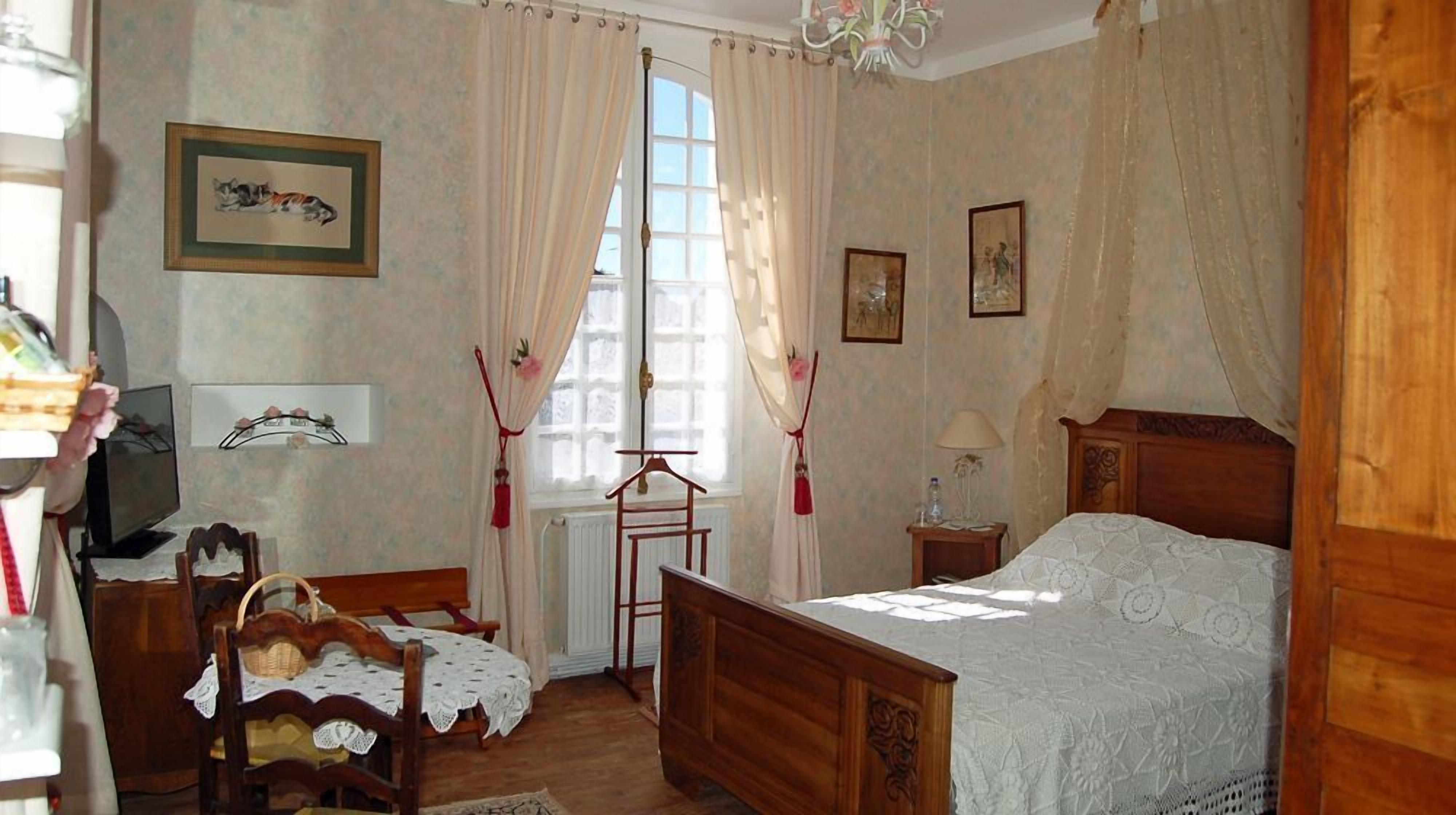A cozy bedroom with a wooden bed, a chair, a small table, and a window with curtains.