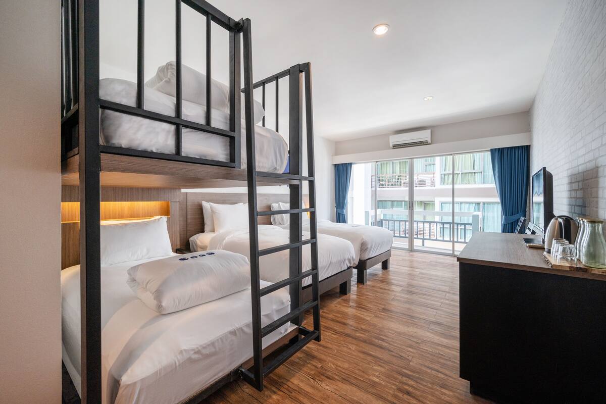 A hotel room with bunk beds, a desk with a kettle, and a view of the city through large windows.