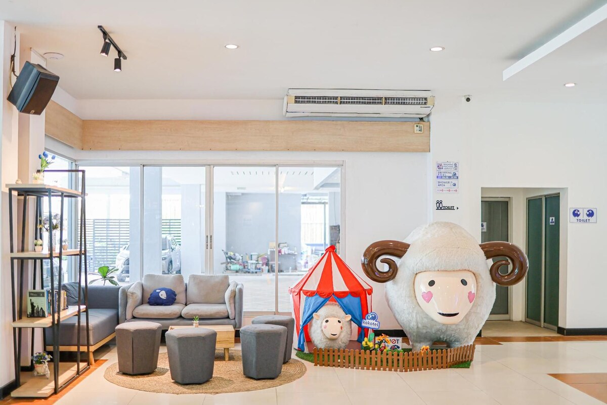 A hotel lobby with a large sheep statue, a sofa, ottomans, a bookshelf, and a view through the glass doors.