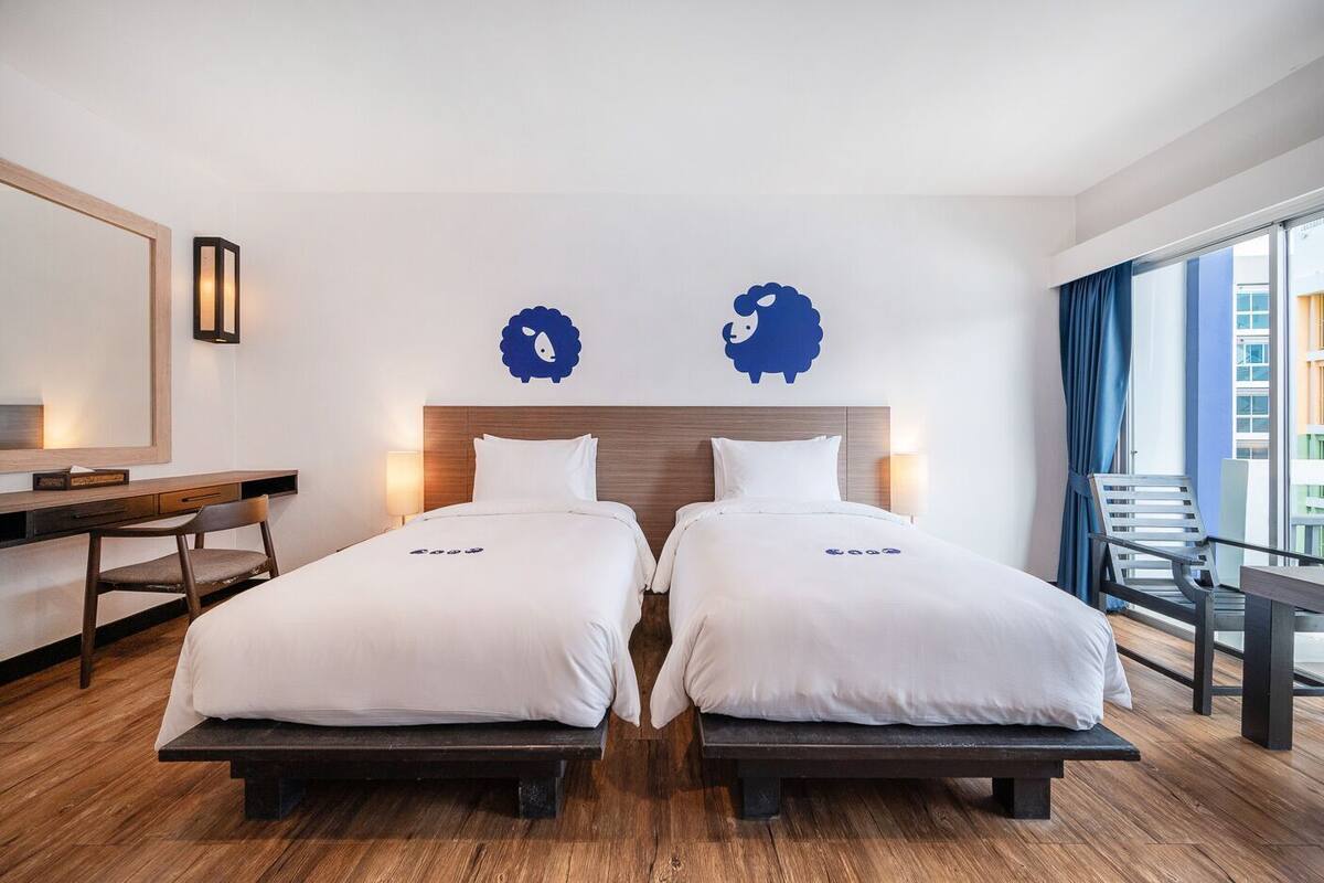 A hotel room with two beds, a desk, a chair, a large window with curtains, and a wall-mounted lamp.