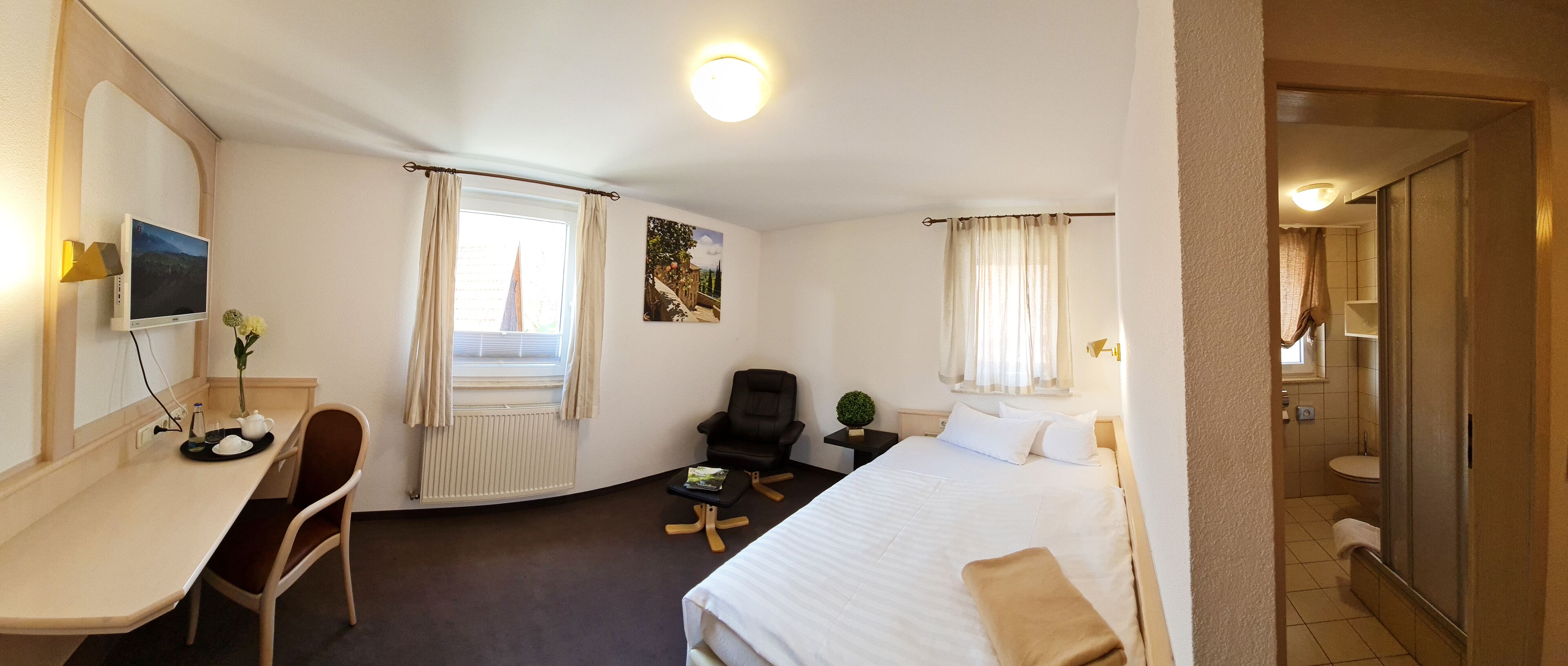 Habitación de hotel con cama, escritorio con silla, televisor, ventana con cortinas, una mesita con planta y baño visible a través de una puerta abierta.
