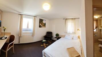 Habitación de hotel con cama, escritorio con silla, televisor, ventana con cortinas, una mesita con planta y baño visible a través de una puerta abierta.
