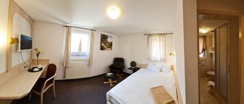 Habitación de hotel con cama, escritorio con silla, televisor, ventana con cortinas, una mesita con planta y baño visible a través de una puerta abierta.