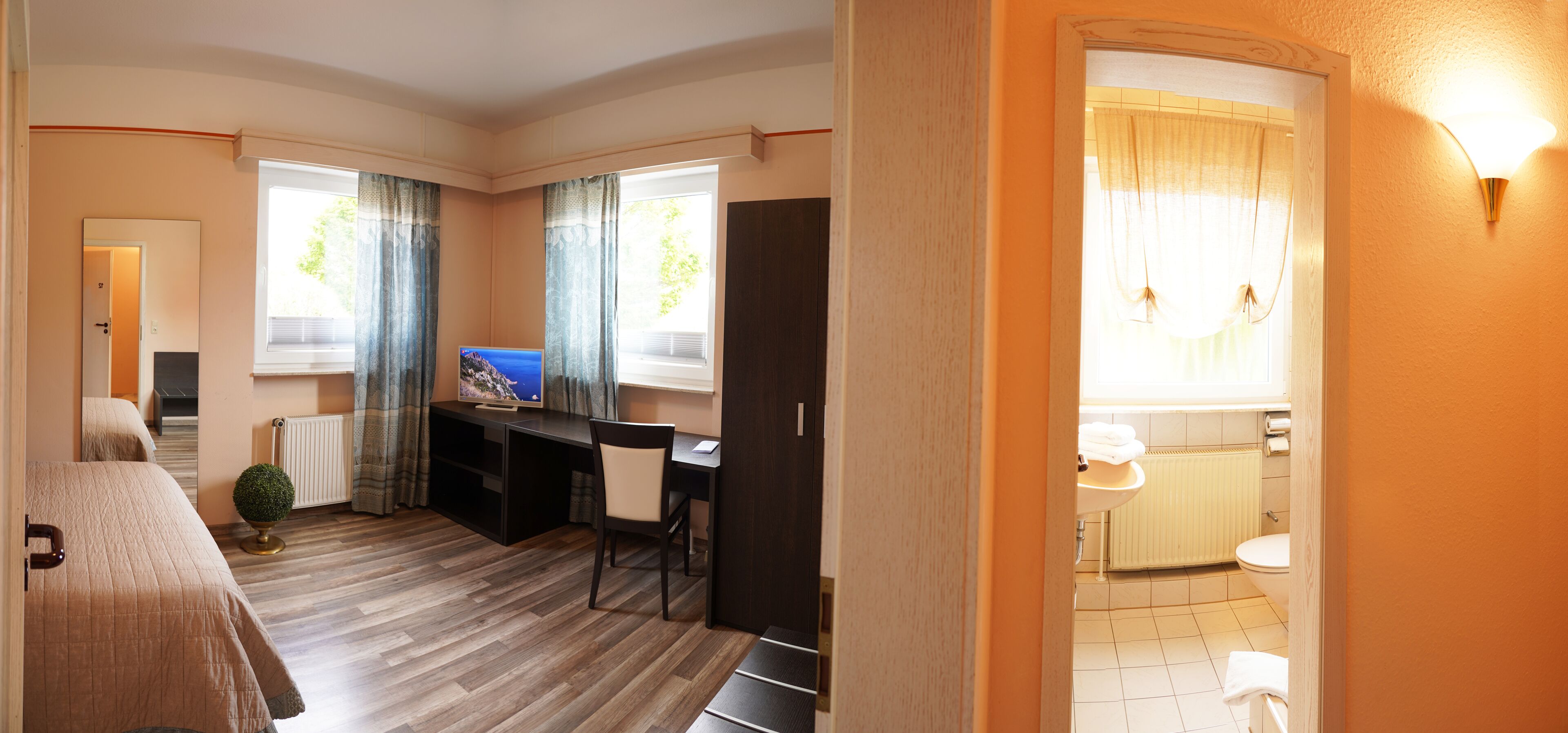 A hotel room with a bed, a desk, a chair, a small plant, and a bathroom with a toilet and sink.