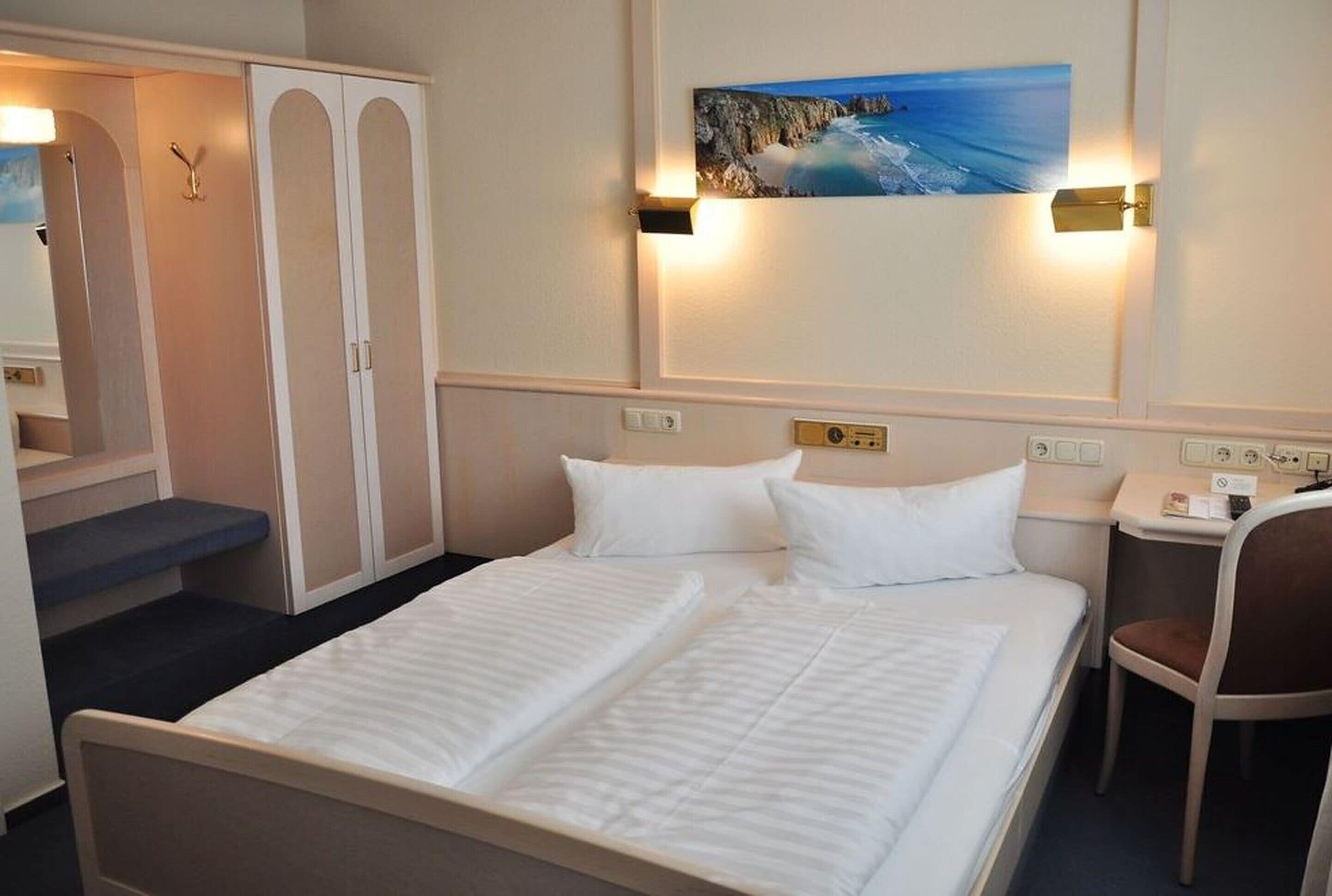 Ein Hotelzimmer mit Bett, Schreibtisch, Stuhl und einem Gemälde einer Küstenlandschaft.
