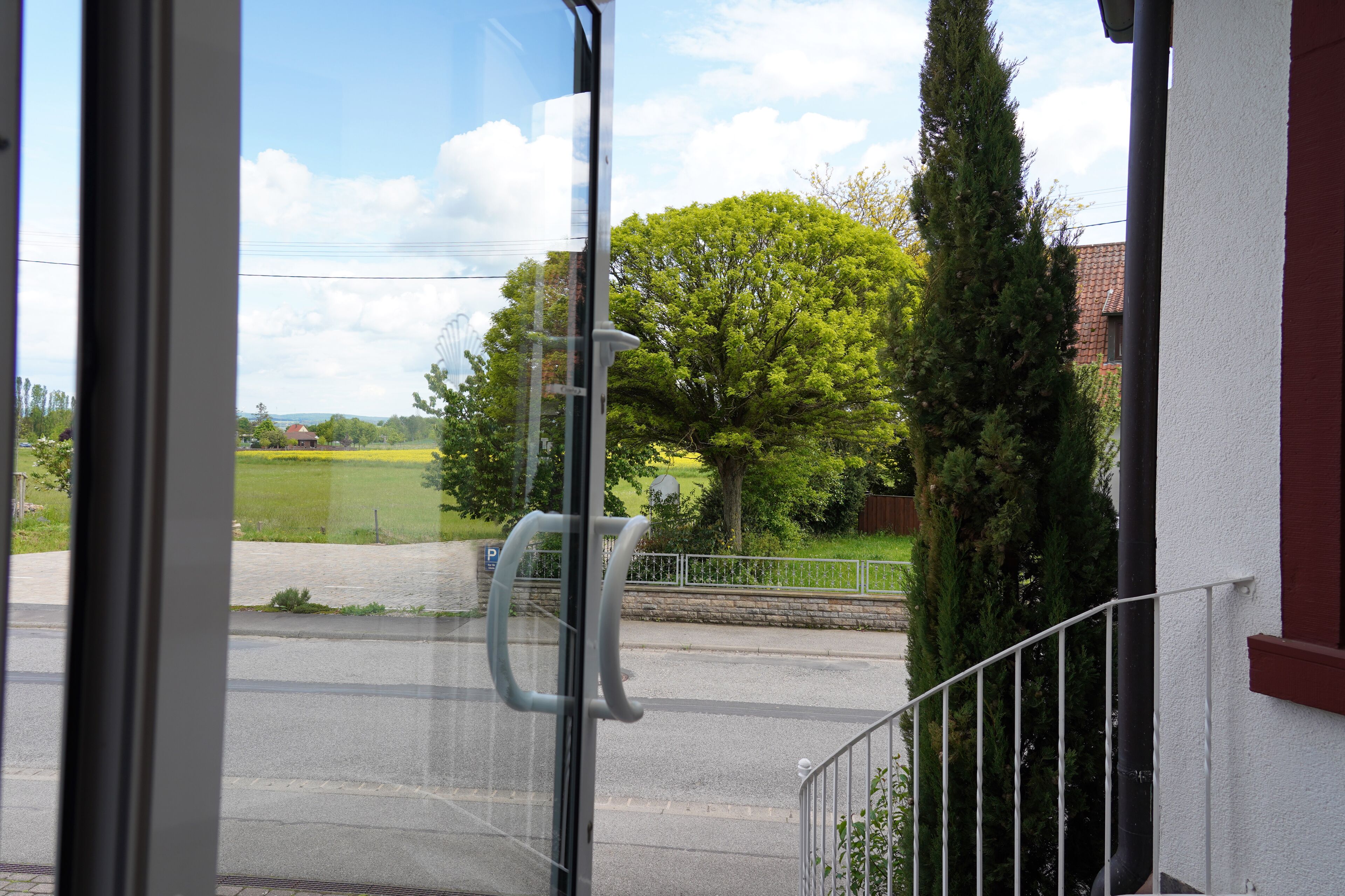 Blick aus einem Fenster mit Metallgeländer, bewölkter Himmel und einer grünen Landschaft mit Bäumen und einem Gebäude im Hintergrund.