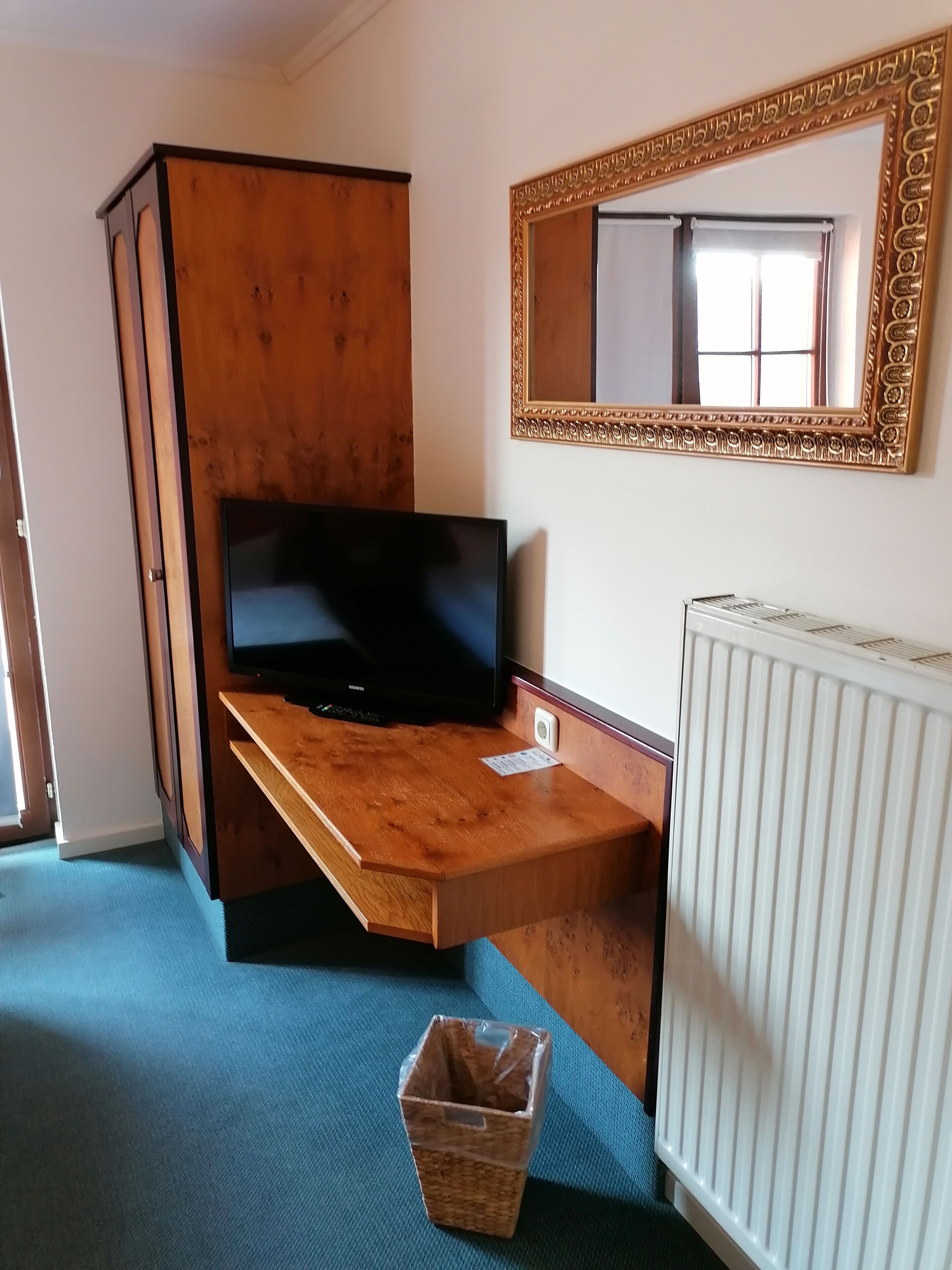 Quarto com escrivaninha de madeira, televisão, radiador, espelho e tapete azul.