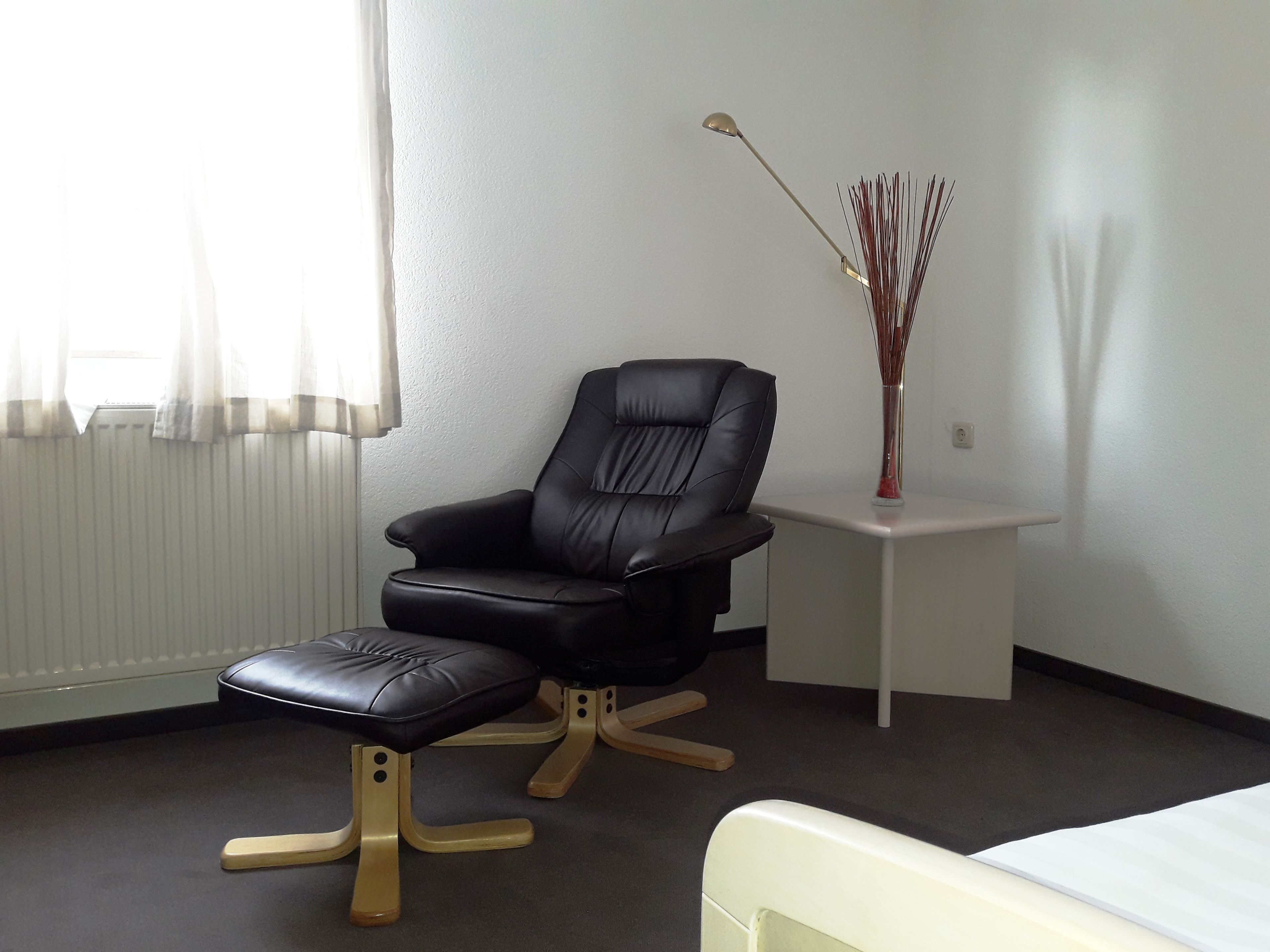 A modern room with a black leather chair, a small white table with a vase, and a white bed.