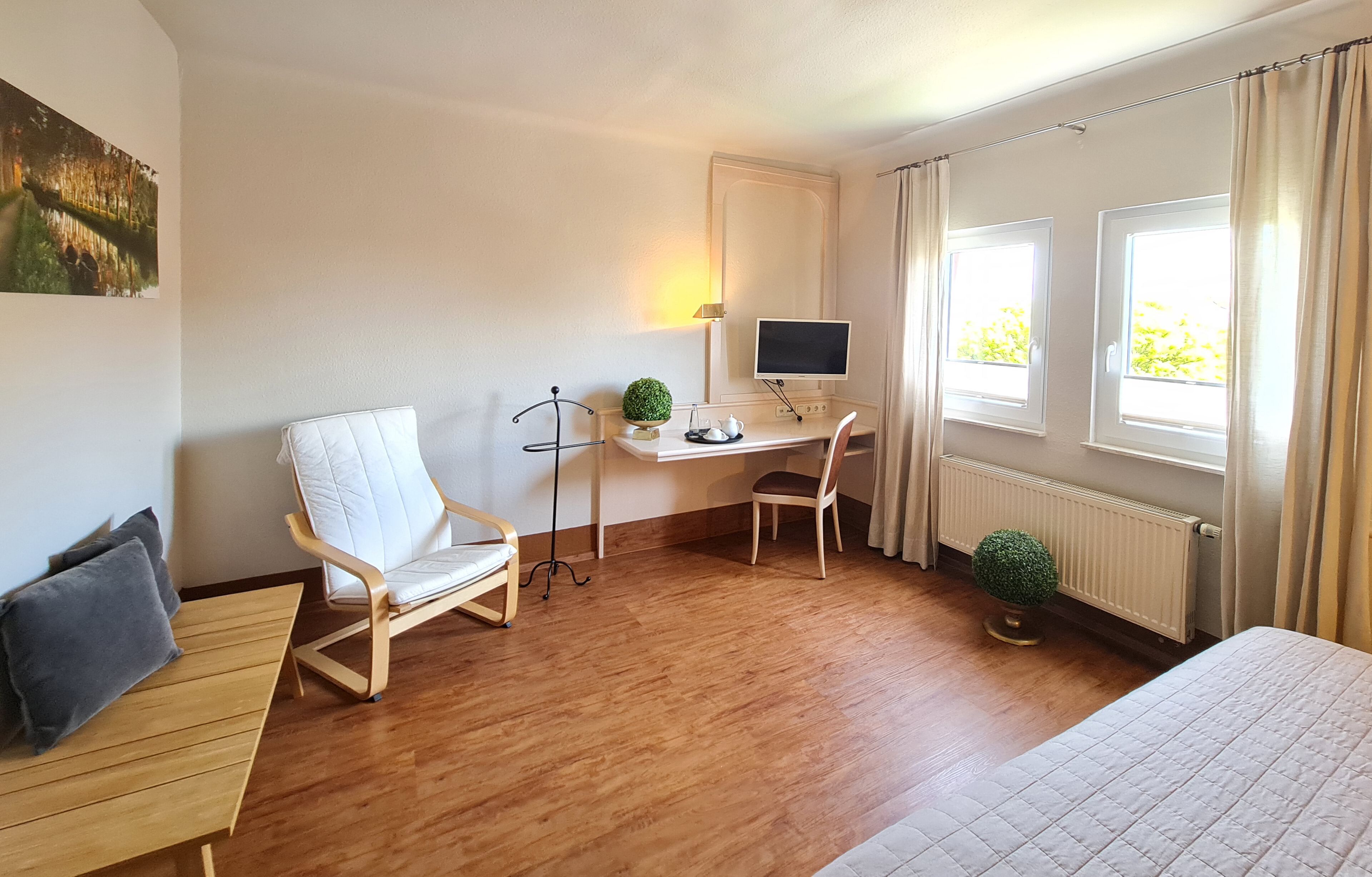 Habitación de hotel con suelo de madera, un escritorio con computadora, una silla, una cama y dos ventanas con cortinas.