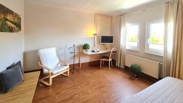 Habitación de hotel con suelo de madera, un escritorio con computadora, una silla, una cama y dos ventanas con cortinas.