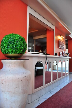 Zona de recepción con un jarrón blanco grande, mostrador de recepción y pared roja.