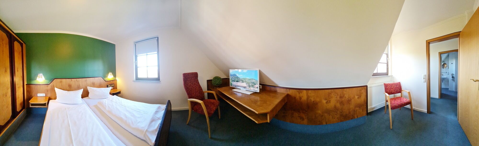 Quarto de hotel com cama, escrivaninha, cadeira e televisão.