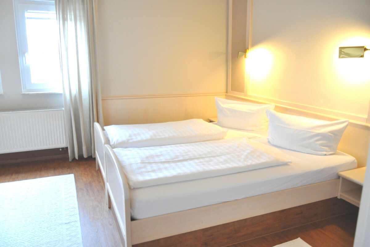 Quarto de hotel com cama de casal, mesa de cabeceira, cortinas na janela e luminária fixa na parede.